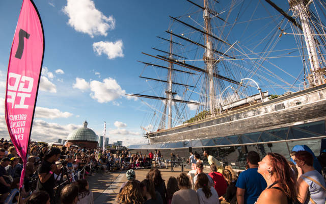 GDIF 2015, Greenwich Fair. Crowds gather round the Cutty Sark and pink flags to watch an outdoor arts show on a sunny London day.