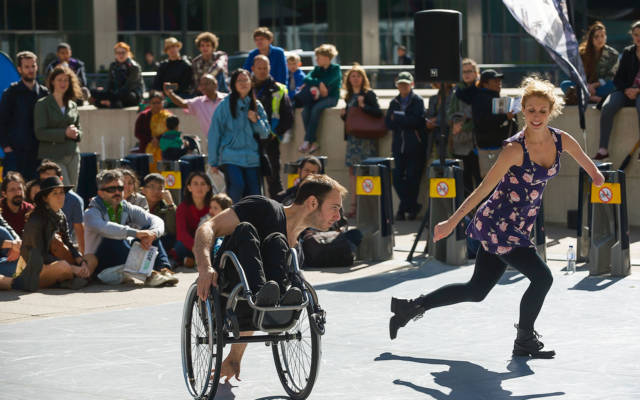 Dedicated To..., Candoco Dance Company, Dancing City, GDIF 2018. A crowd watches a dancer in a wheelchair and a dancer with one arm in Canary Wharf.