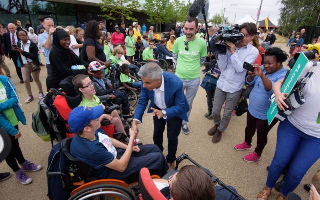 Sadiq Khan shakes hands with a man in a wheelchair, surrounded by crowds, young people in wheelchairs, and a TV camera. Festival.org.