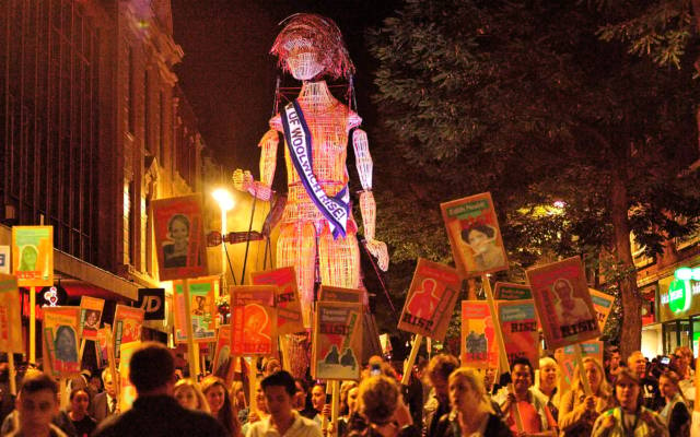 Women of Woolwich Rise, L'Homme Debout and Festival.org, GDIF 2018. A giant puppet wearing a suffragette sash is surrounded by people carrying placards in Woolwich.