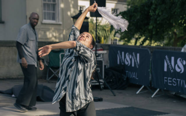 A tall, human sized puppet with a pale wooden face and arm, draped in a white gown. There are 2 puppeteers either side. One is holding it’s head up and arm reaching outward. The other is knelt in a lunge placing a red feather in it’s hands.