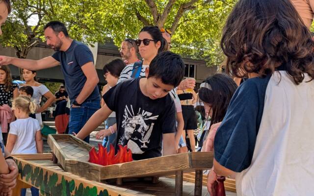 Children and families gathered around a wooden game, shaped like a table top in the sunshine.