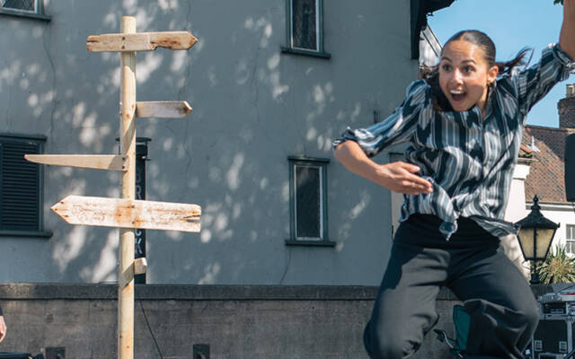 A woman joyfully leaping in in the air with one arm in the air and an open grin on her face. On the left side of the image there is a wooden sign post, with 4 wooden signs pointing in different directions. At the bottom of the signpost there is a light coloured wooden bowl with a red and black mask in it with black feathers.