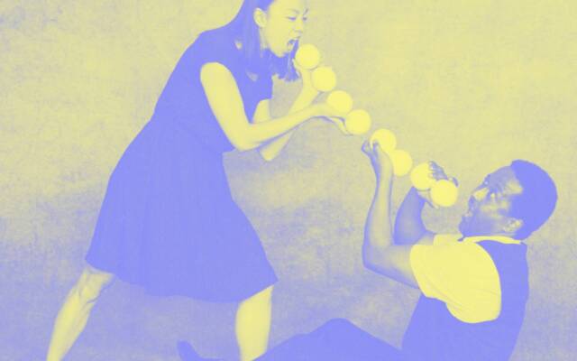 A woman stood facing a man sat on the floor. Thold juggling balls In a chain between each others’ open mouths. The image is edited purple and yellow