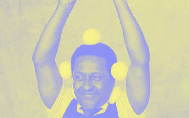 A man holds 2 juggling balls in the air above his head. There are 5 more balls balanced around his head and neck. He is smiling. The image is edited purple and yellow