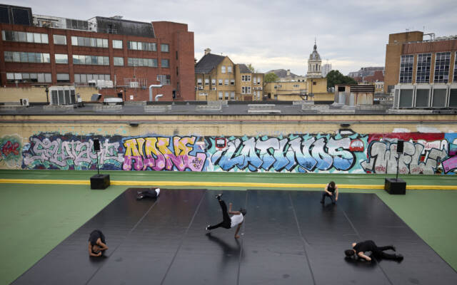 A wide angle image of a city scene, four performers are performing on a matt. Behind them is a wall with colourful graffiti.