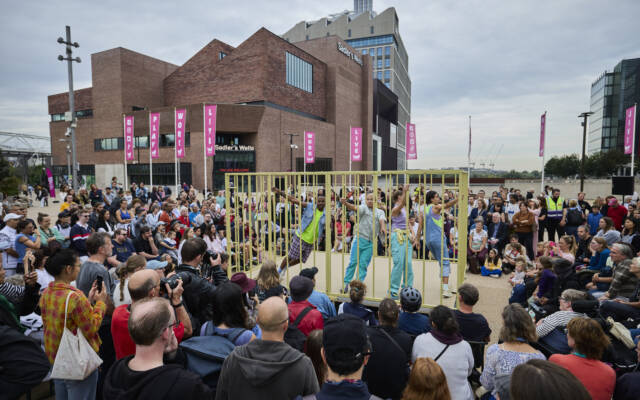 A busy street scene. A large crowd surrounding a yellow cage with a solo performer inside.