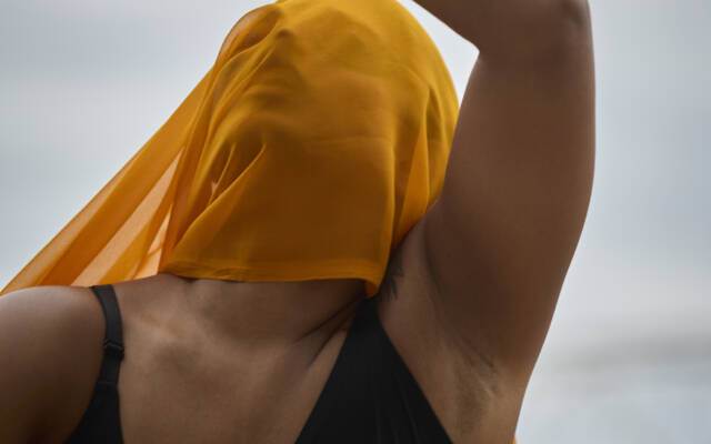 Woman wearing an orange scarf over her face. Her arm is raised above her head. Behind her is a dark overcast sky