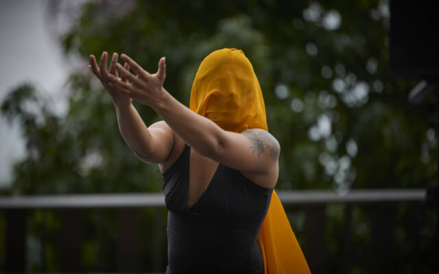 Woman wearing an orange scarf over her face. Her arms are stretched out in front of her.